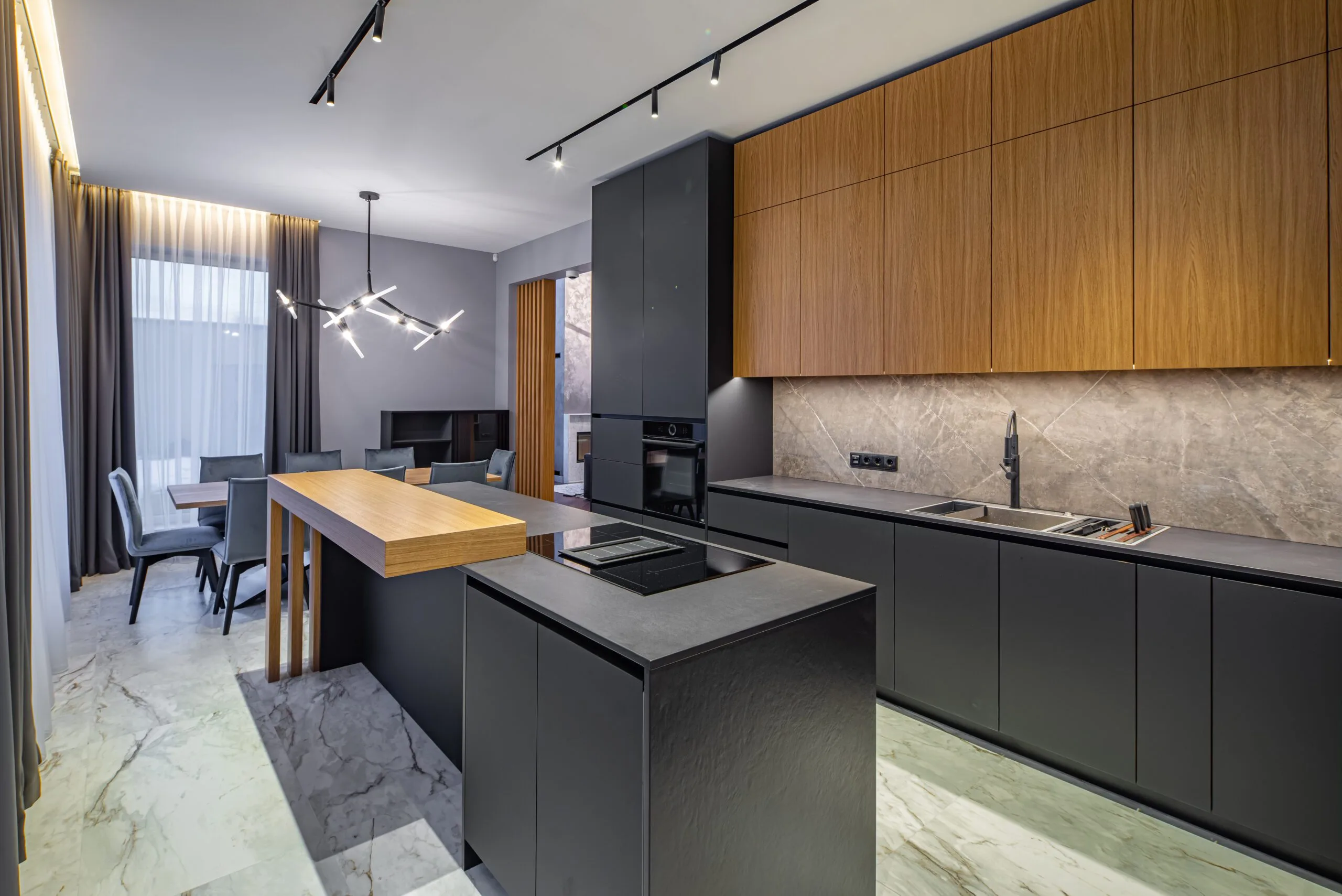 Modern kitchen with black cabinets, marble countertops, a wooden island, and an industrial-style light fixture. Dining area with chairs and a table in the background.